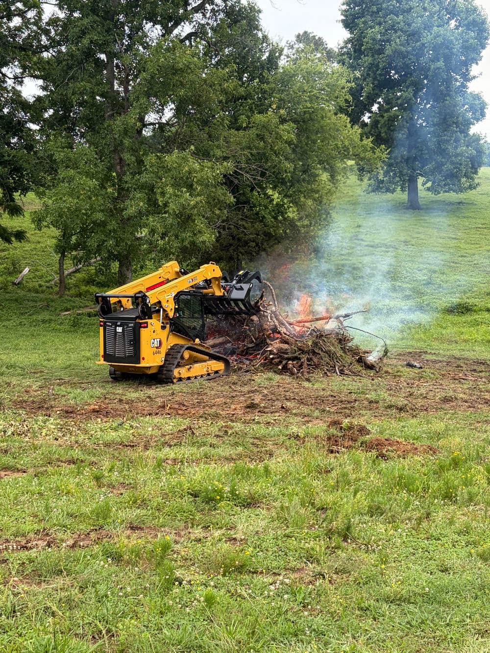 Caterpillar mini excavator managing a controlled burn of tree branches in a grassy field.