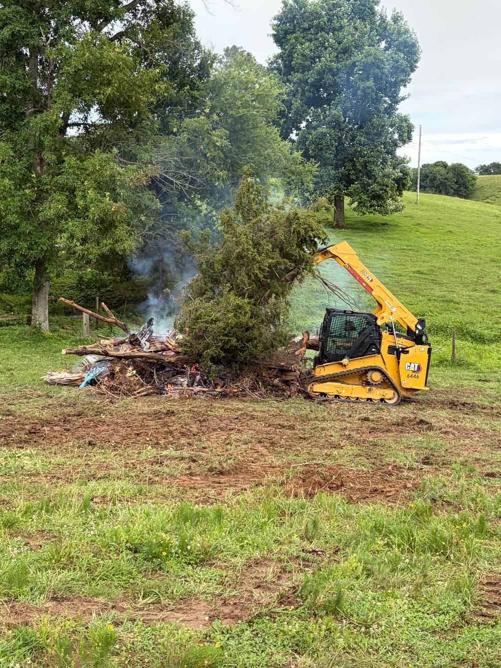 Skid steer moving brush pile with smoke on a grassy landscape.
